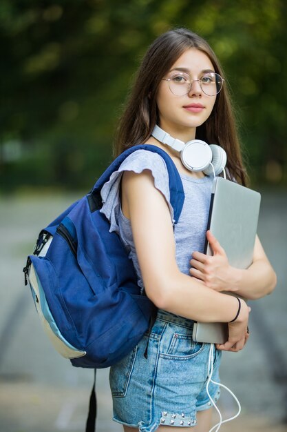 Smiling young girl student with backpack holding mobile phone, walking at the park, listening to music with earphones