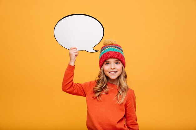 Smiling Young girl in sweater and hat holding blank speech buble and looking at the camera over orange