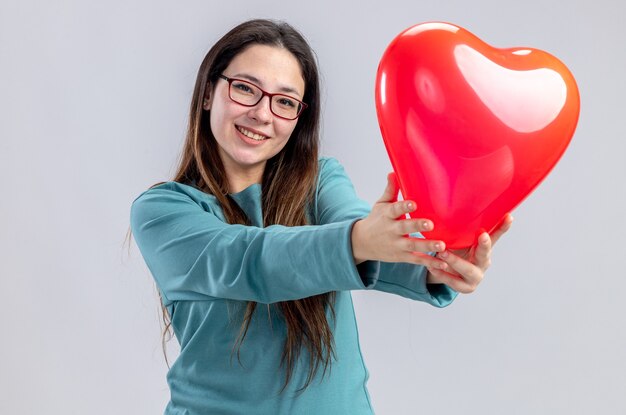 Smiling young girl on valentines day holding out heart balloon at camera isolated on white background