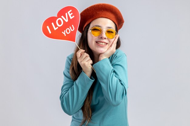 Smiling young girl on valentines day wearing hat with glasses holding red heart on a stick with i love you text putting hand on cheek isolated on white background