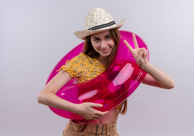 Smiling young girl wearing hat and swim ring doing peace sign on isolated white wall