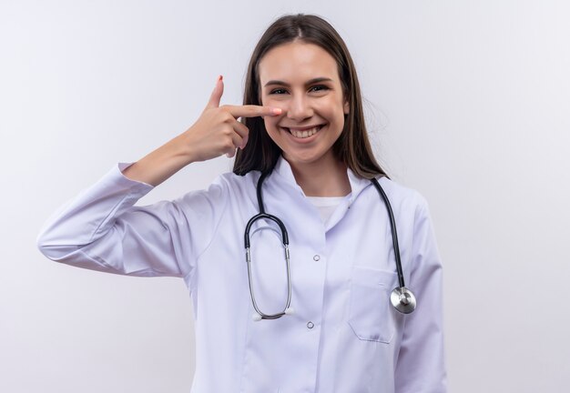 Smiling young girl wearing stethoscope medical gown points finger to nose on isolated white background