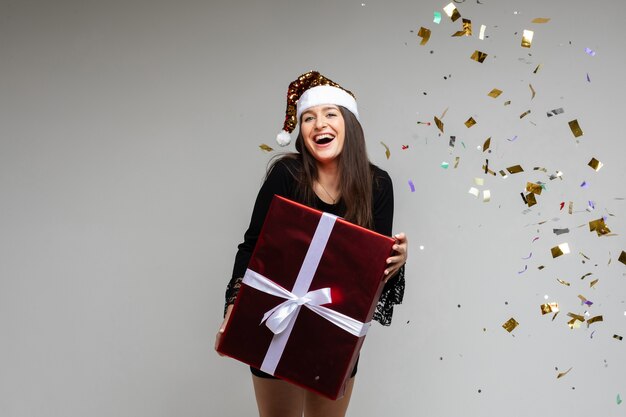 Smiling young girl with large festive gift pointing by hand on empty space with holiday confetti on gray background