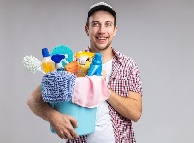 Smiling young guy cleaner wearing cap holding bucket with cleaning tools isolated on white background