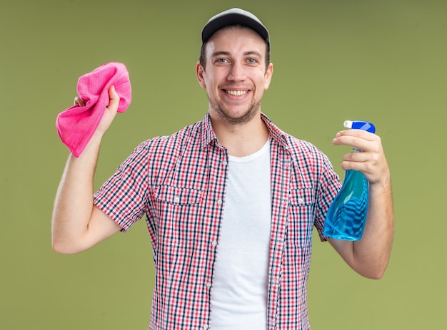 Улыбающийся молодой парень уборщик в кепке держит чистящее средство с тряпкой на оливково-зеленом фоне