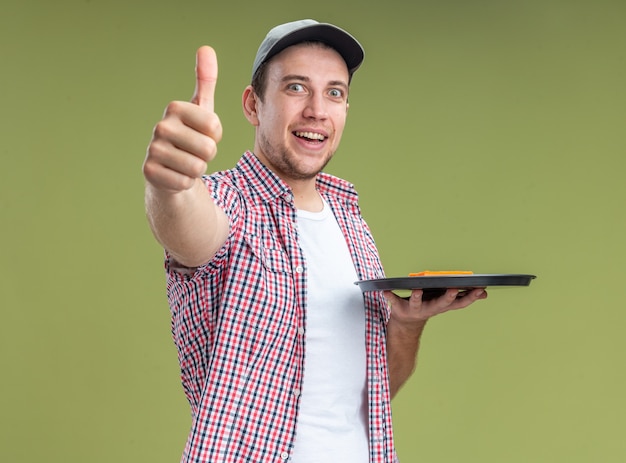 Улыбающийся молодой парень уборщик в кепке держит губку на подносе, показывая большой палец вверх, изолированные на оливково-зеленом фоне