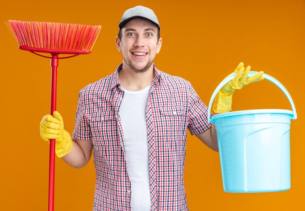 Smiling young guy cleaner wearing cap with gloves holding bucket and mop isolated on orange wall