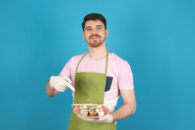 Smiling young guy holding cake slices and pointing finger at it.