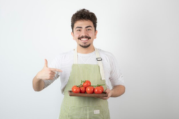 Smiling young guy holding pile of organic tomatoes and pointing finger on it.