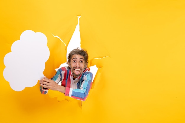 Free photo smiling young guy holding white cloud-shaped paper and posing for camera in a torn hole and free background in yellow paper