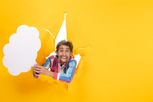 Улыбающийся молодой парень держит белую бумагу в форме облака и позирует перед камерой в разорванной дыре на свободном фоне в желтой бумаге