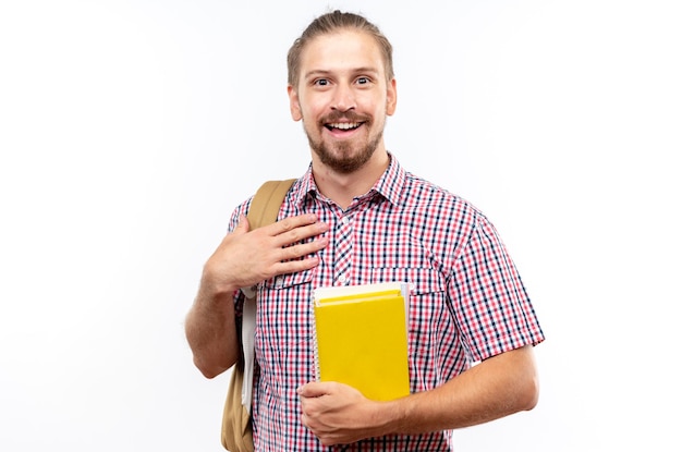 Smiling young guy student wearing backpack holding books putting hand on himself isolated on white wall
