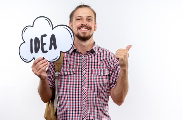 Smiling young guy student wearing backpack holding idea bubble showing thumb up isolated on white wall