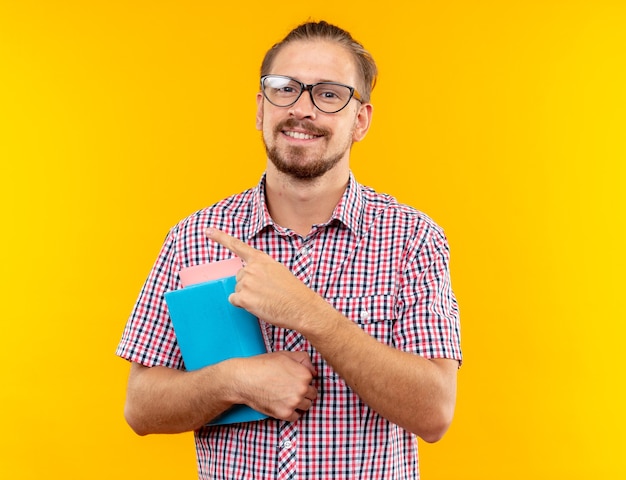 Улыбающийся молодой парень-студент в рюкзаке с очками, держащий книжные очки сбоку, изолирован на оранжевой стене с копией пространства