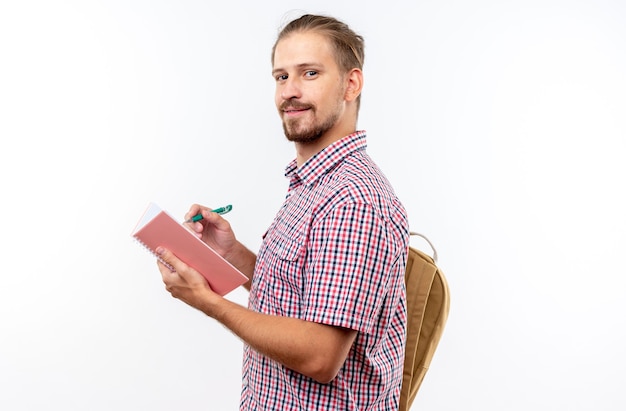 白い壁で隔離のノートに何かを書くバックパックを身に着けている若い男の学生の笑顔
