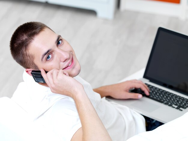 Smiling young guy using mobile phone and laptop at home