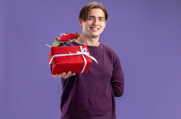Улыбающийся молодой парень в день святого валентина протягивает подарочную коробку перед камерой, изолированной на синем фоне