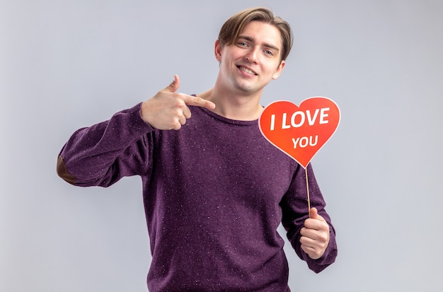 Sorridente giovane ragazzo il giorno di san valentino che tiene e punta al cuore rosso su un bastone con ti amo testo isolato su sfondo bianco