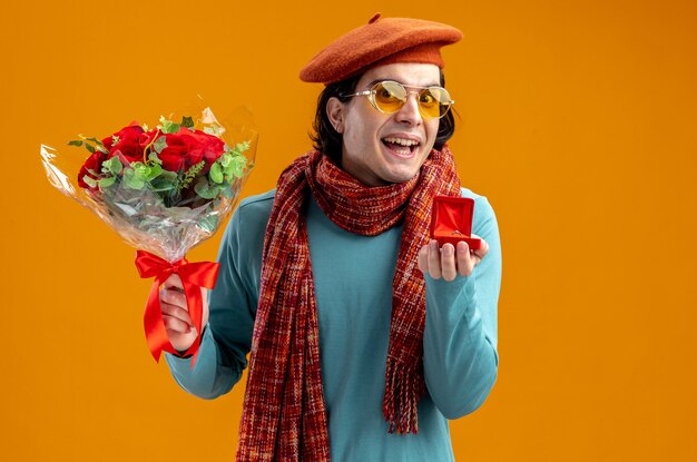 Smiling young guy on valentines day wearing hat with scarf and glasses holding bouquet with wedding ring isolated on orange background