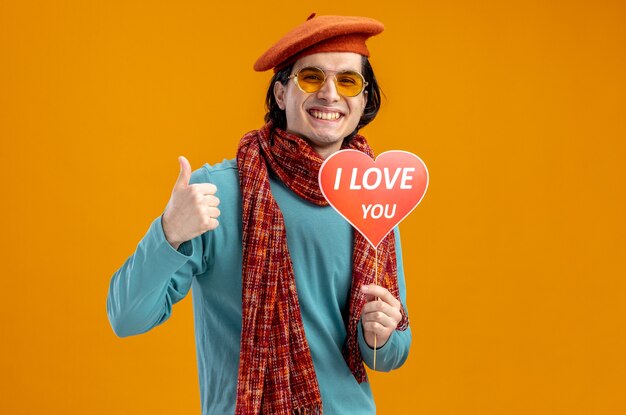 발렌타인 데이에 웃고 있는 젊은 남자가 스카프와 안경을 쓴 채 막대기에 빨간 하트를 들고 있고 주황색 배경에 고립된 엄지손가락을 보여주는 텍스트를 사랑합니다.