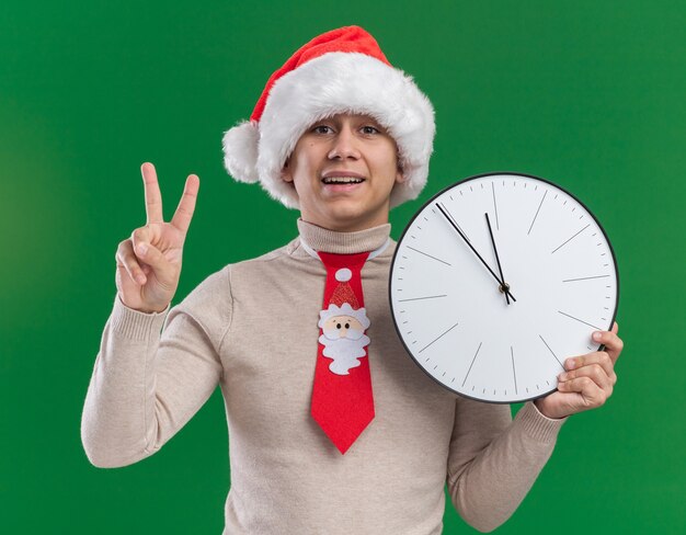 Smiling young guy wearing christmas hat with tie holding wall clock showing peace gesture isolated on green wall