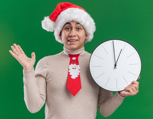 Smiling young guy wearing christmas hat with tie holding wall clock spreading hand isolated on green wall
