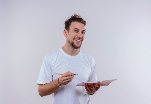 孤立した白い背景に鉛筆とノートブックを保持している白いTシャツを着て笑顔の若い男