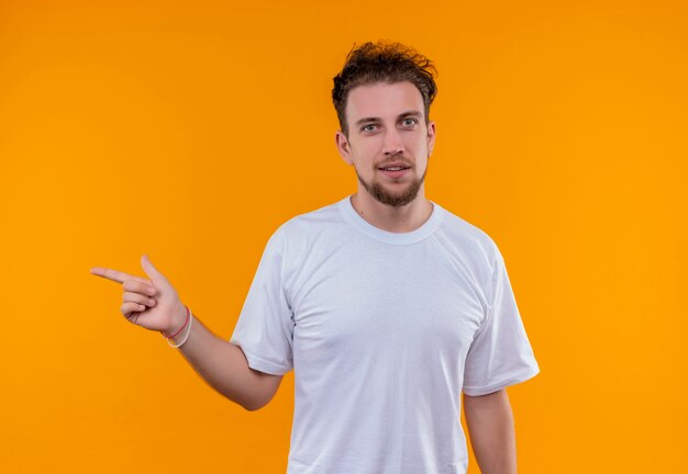 Smiling young guy wearing white t-shirt points to side on isolated orange background