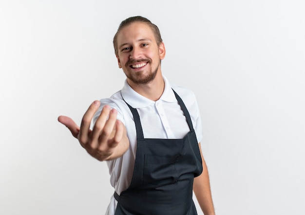 Foto gratuita sorridente giovane barbiere bello indossando l'uniforme facendo venire qui gesto davanti isolato sul muro bianco