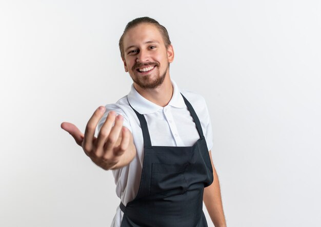 Smiling young handsome barber wearing uniform doing come here gesture at front isolated on white wall