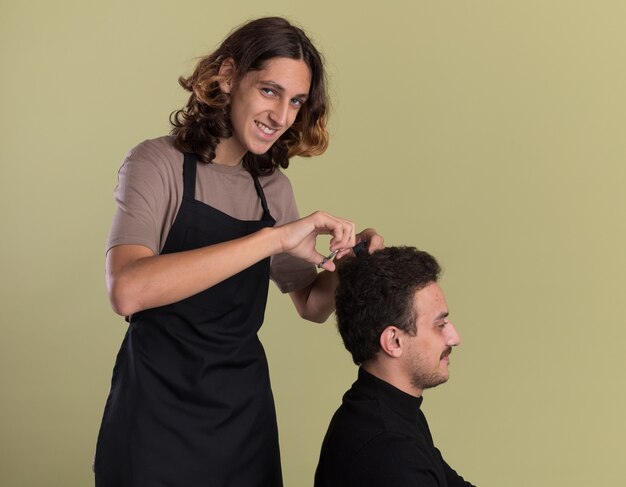 Smiling young handsome barber wearing uniform doing haircut for his young client