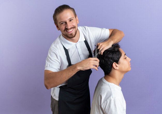Smiling young handsome barber wearing uniform doing haircut for young client isolated on purple wall
