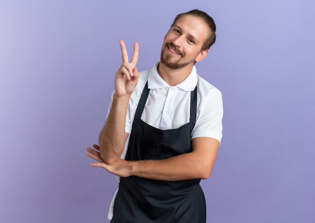 Smiling young handsome barber wearing uniform doing peace sign and putting hand under elbow isolated on purple wall