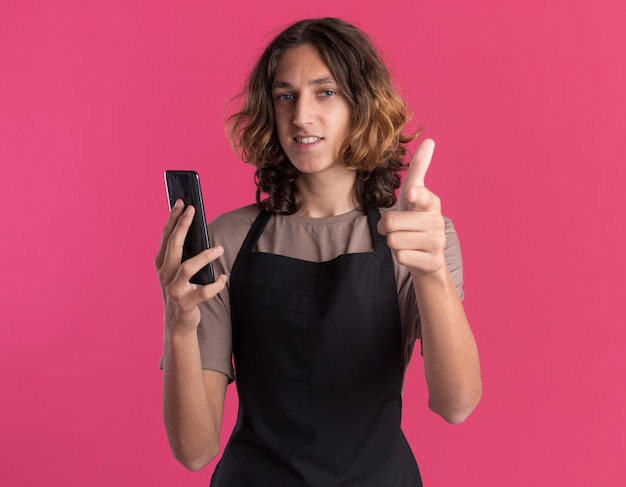 Улыбающийся молодой красивый парикмахер в униформе держит мобильный телефон, глядя и указывая на розовую стену с копией пространства