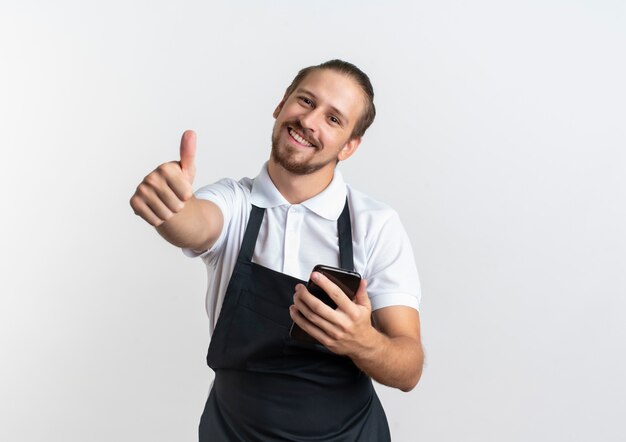 Smiling young handsome barber wearing uniform holding mobile phone and showing thumb up isolated on white wall