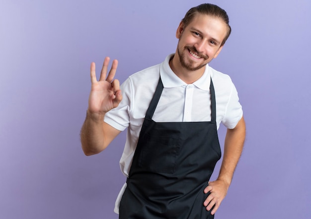 Smiling young handsome barber wearing uniform putting hand on waist and doing ok sign isolated on purple wall