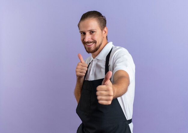 Smiling young handsome barber wearing uniform showing thumbs up at front isolated on purple wall