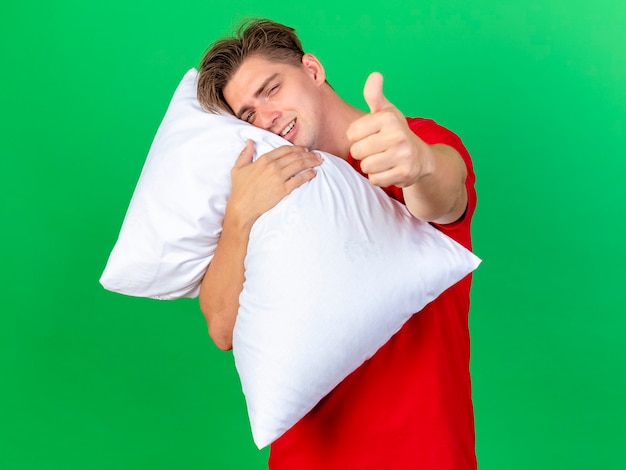 Улыбающийся молодой красивый блондин больной мужчина держит подушку, положив голову на подушку, глядя в камеру, показывая большой палец вверх, изолированный на зеленом фоне с копией пространства