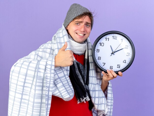 Smiling young handsome blonde ill man wearing winter hat and scarf wrapped in plaid holding clock looking at camera showing thumb up isolated on purple background