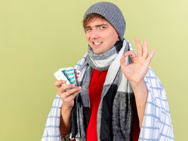 Smiling young handsome blonde ill man wearing winter hat and scarf wrapped in plaid holding packs of medical pills  doing ok sign isolated on olive green wall with copy space