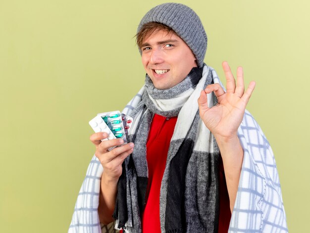 Smiling young handsome blonde ill man wearing winter hat and scarf wrapped in plaid holding packs of medical pills  doing ok sign isolated on olive green wall with copy space