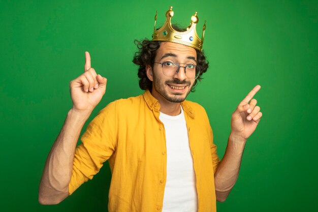 Smiling young handsome caucasian man wearing glasses and crown  pointing up isolated on green wall
