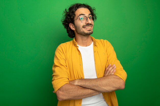 Free photo smiling young handsome caucasian man wearing glasses standing with closed posture looking at camera isolated on green background with copy space