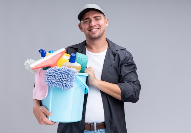 Smiling young handsome cleaning guy wearing t-shirt and cap holding bucket of cleaning tools isolated on white wall with copy space