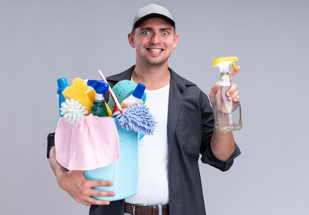 Smiling young handsome cleaning guy wearing t-shirt and cap holding bucket of cleaning tools and spray bottle isolated on white wall