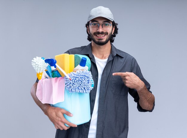 Smiling young handsome cleaning guy wearing t-shirt and cap holding and points at bucket of cleaning tools isolated on white wall