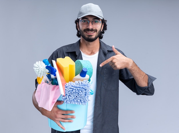 Free photo smiling young handsome cleaning guy wearing t-shirt and cap holding and points at bucket of cleaning tools isolated on white wall