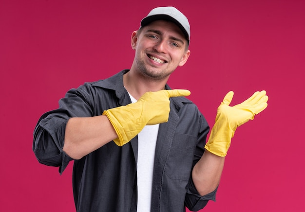 Улыбающийся молодой красивый парень-уборщик в футболке и кепке с перчатками притворяется, что держит, и указывает на что-то изолированное на розовой стене
