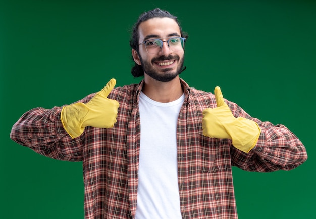 Sorridente giovane bel ragazzo delle pulizie indossando t-shirt e guanti che mostrano i pollici in su isolato sulla parete verde