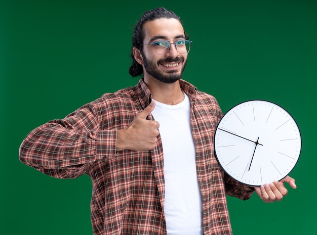 Smiling young handsome cleaning guy wearing t-shirt holding wall clock showing thumb up isolated on green wall